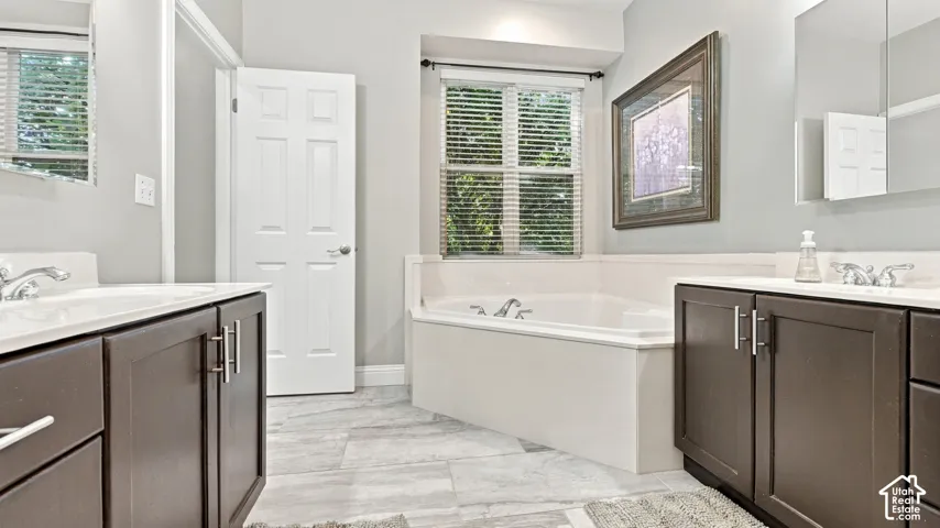 Dual vanities featuring dark cabinetry, solid-surface countertops, and undermount sinks, accented by a deep soaking tub with a tiled surround and large-format tile flooring