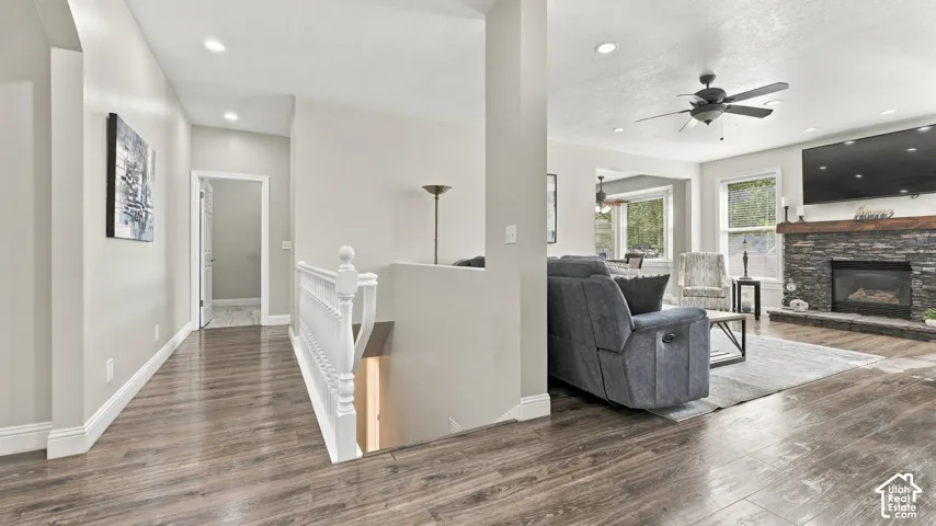 Wood-finish flooring, stone-surround fireplace with a timber mantel, white balustrade staircase railing, recessed lighting, and ceiling fan