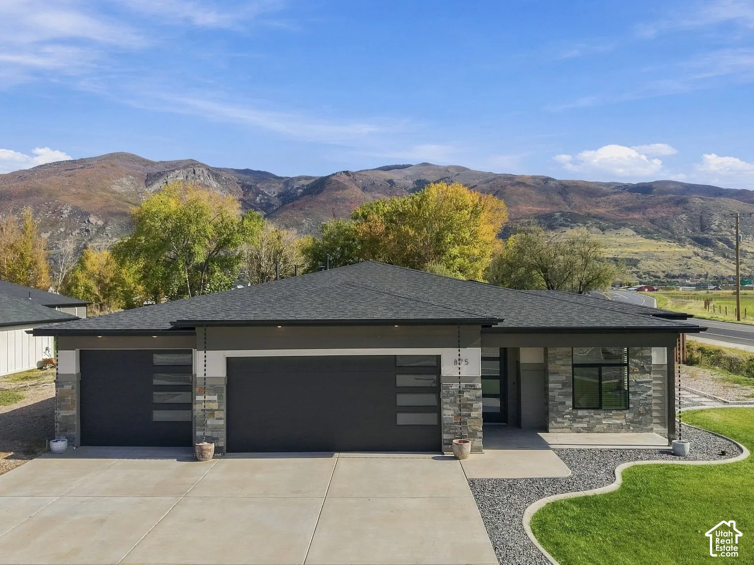 Single-story exterior featuring multi-tonal stone veneer, horizontal panel siding, a concrete driveway, two-car garage with glass-paneled doors, and mountain backdrop