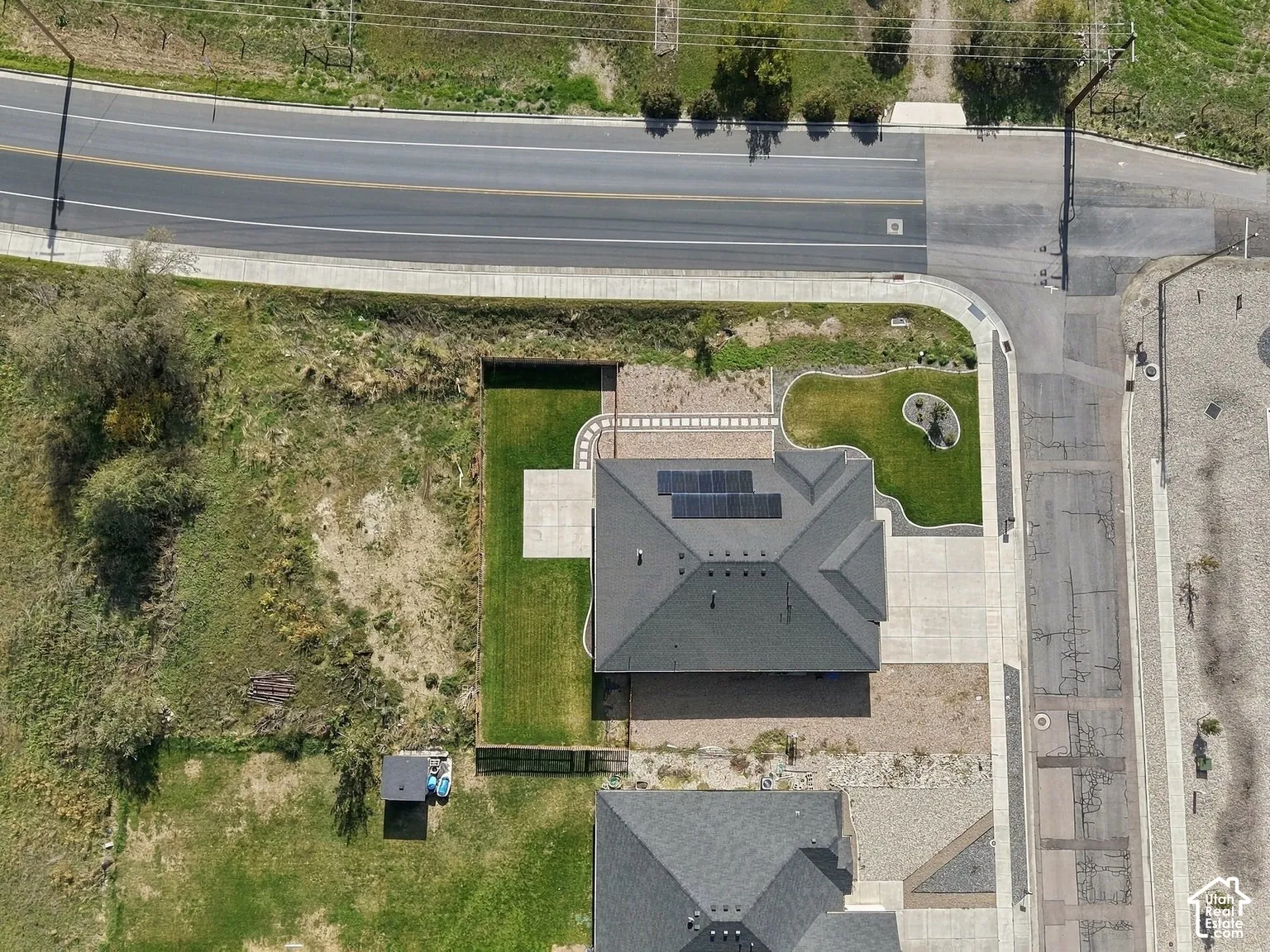 Corner lot featuring a concrete driveway, roof-mounted solar panels, paved walkway, manicured lawn, and hardscaped patio area