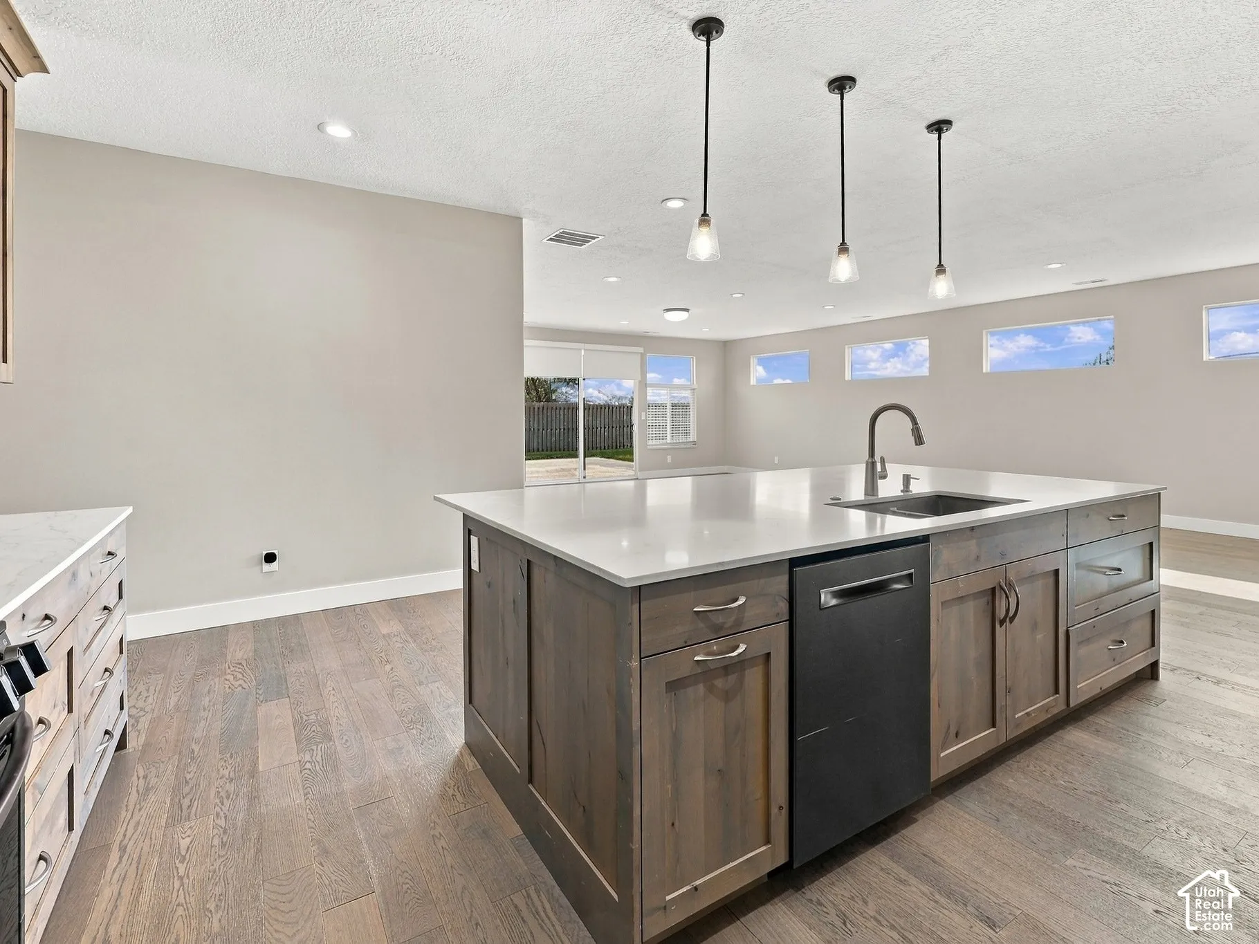 Kitchen featuring an expansive center island with stone-surface countertop, dark wood-finish cabinetry, built-in dishwasher, and three pendant lights, complemented by wood-finish flooring and recessed lighting
