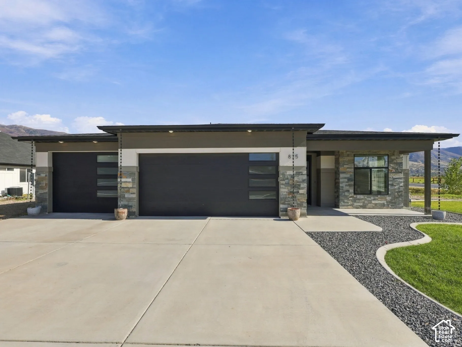 Modern single-story exterior featuring multi-tonal stone veneer, horizontal panel siding, dark-finish garage doors with frosted glass inserts, and decorative rain chains