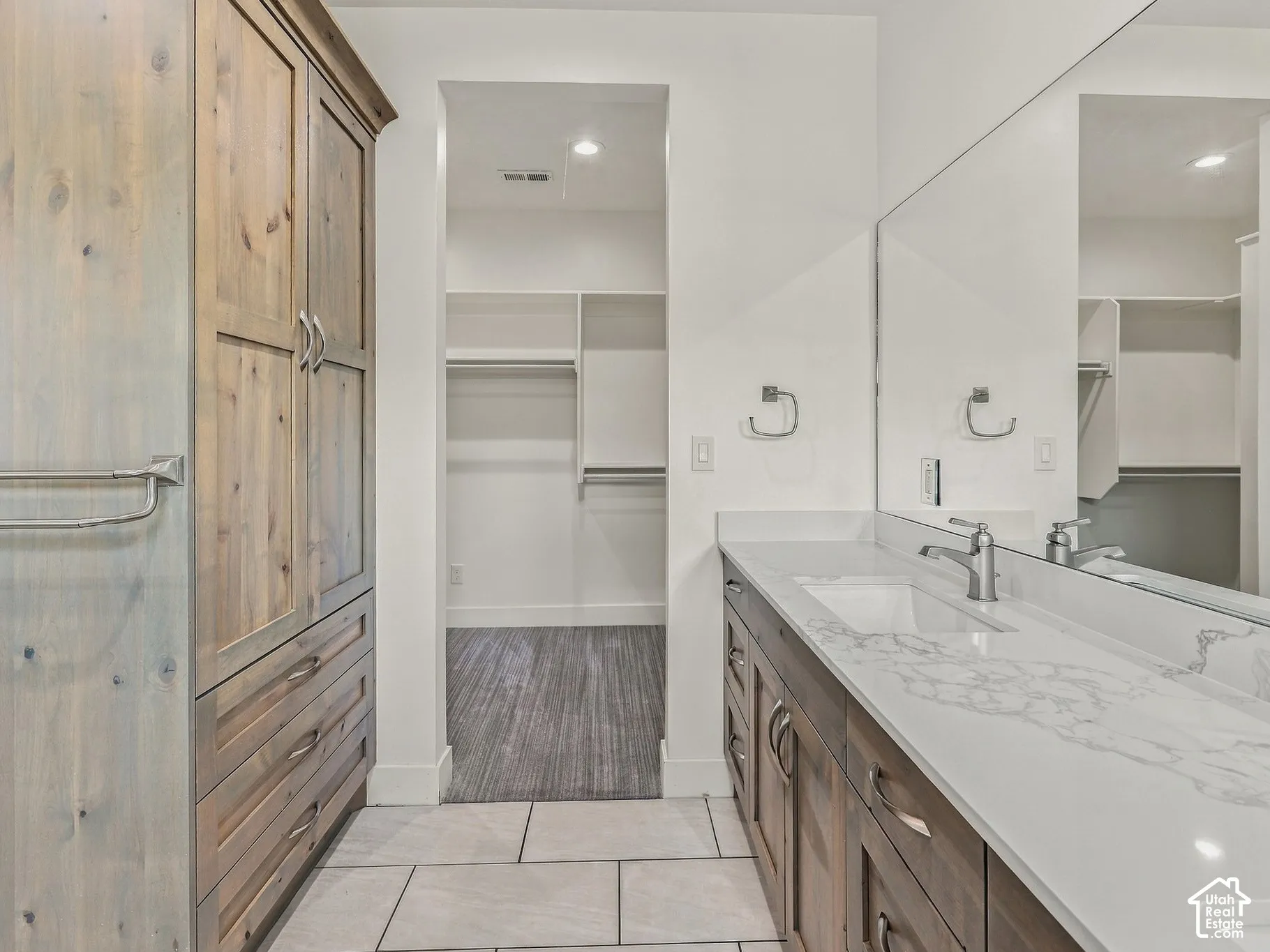 Bathroom featuring wood-finish cabinetry, stone-patterned countertop with integrated sink, floor-to-ceiling storage hutch, full-width wall mirror, and tile flooring