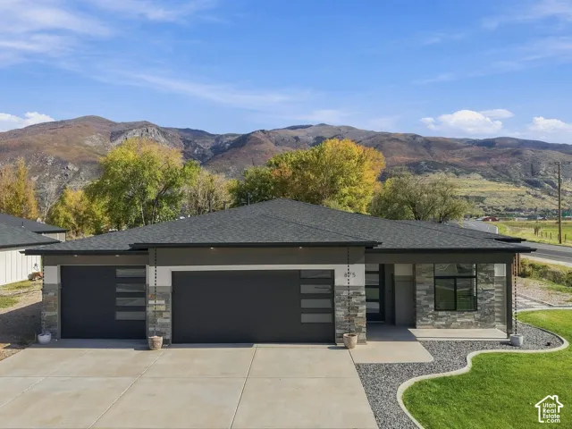 Single-story exterior featuring multi-tonal stone veneer, horizontal panel siding, a concrete driveway, two-car garage with glass-paneled doors, and mountain backdrop