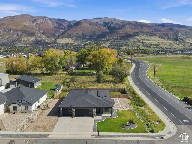 Aerial perspective featuring a single-story residence with a dark metal roof, solar panel array, attached two-car garage, and manicured front lawn set against a mountainous backdrop