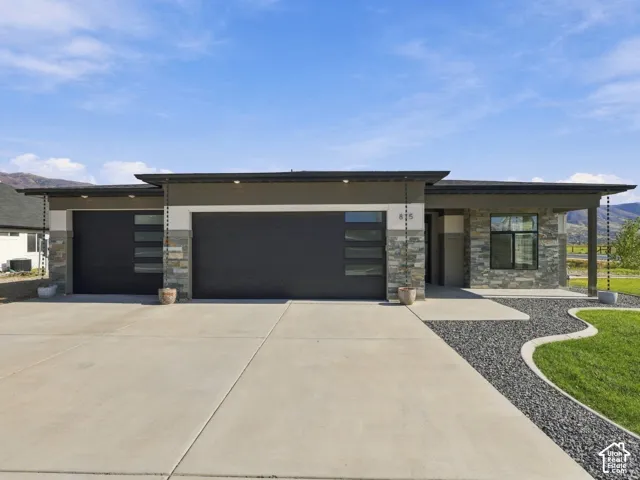 Modern single-story exterior featuring multi-tonal stone veneer, horizontal panel siding, dark-finish garage doors with frosted glass inserts, and decorative rain chains