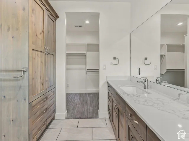 Bathroom featuring wood-finish cabinetry, stone-patterned countertop with integrated sink, floor-to-ceiling storage hutch, full-width wall mirror, and tile flooring