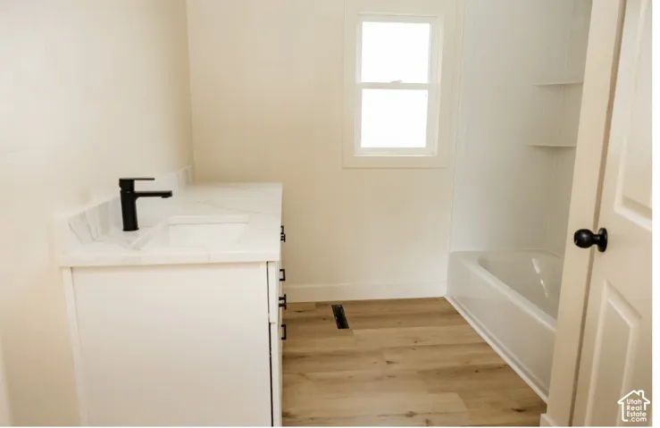 Bathroom featuring a white vanity with a stone-finish countertop, integrated sink, matte black faucet, and wood-finish flooring, complemented by a white bathtub and shower combination