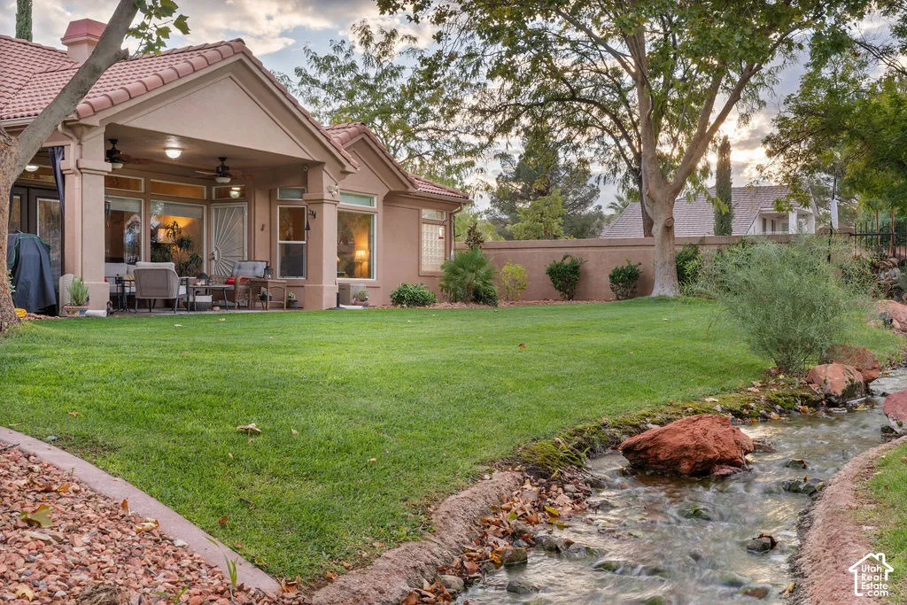 Covered patio featuring recessed lighting and ceiling fans, expansive lawn, natural creek water feature with rock accents, and perimeter masonry wall