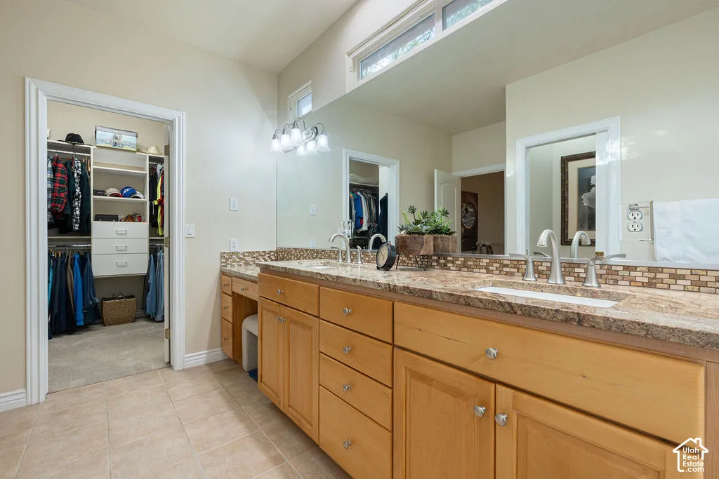Dual-sink vanity with natural stone countertop, tiled backsplash, light wood cabinetry, and large-format wall mirror
