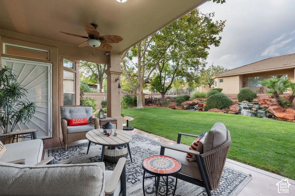 Covered concrete patio featuring a ceiling fan, manicured lawn, tiered rock landscaping, and a built-in water feature