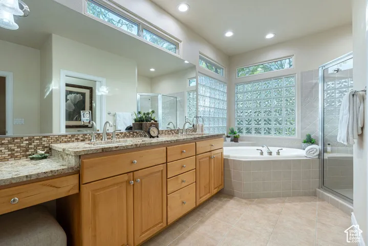 Dual-sink vanity with natural stone countertops, tile backsplash, built-in soaking tub, glass block window detailing, and glass-enclosed shower