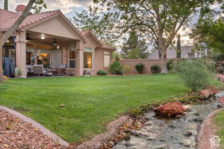 Covered patio featuring recessed lighting and ceiling fans, expansive lawn, natural creek water feature with rock accents, and perimeter masonry wall