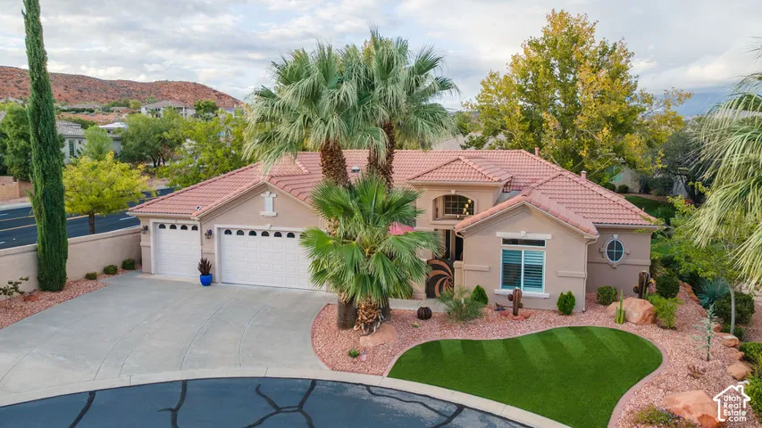 Stucco exterior with a tiled roof, three-car garage, manicured synthetic lawn, and mature palm trees set against a mountain backdrop