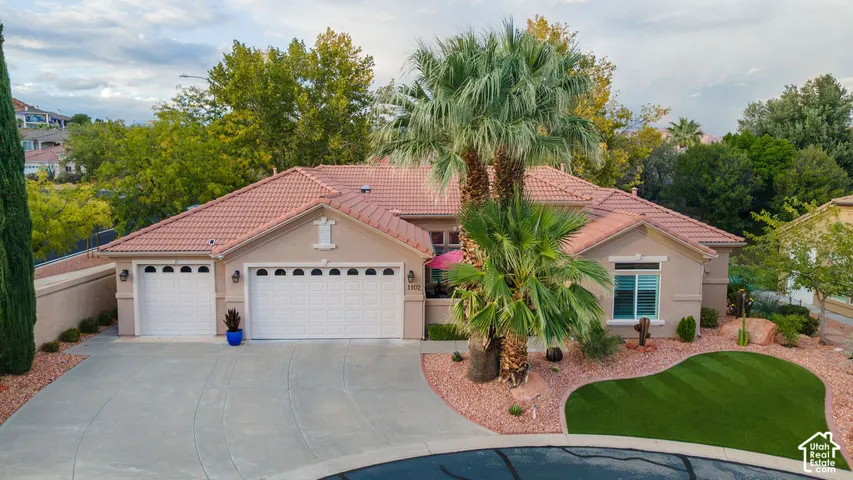 Single-story exterior featuring a clay tile roof, arched garage doors, decorative stucco finishes, mature palm trees, and manicured turf landscaping