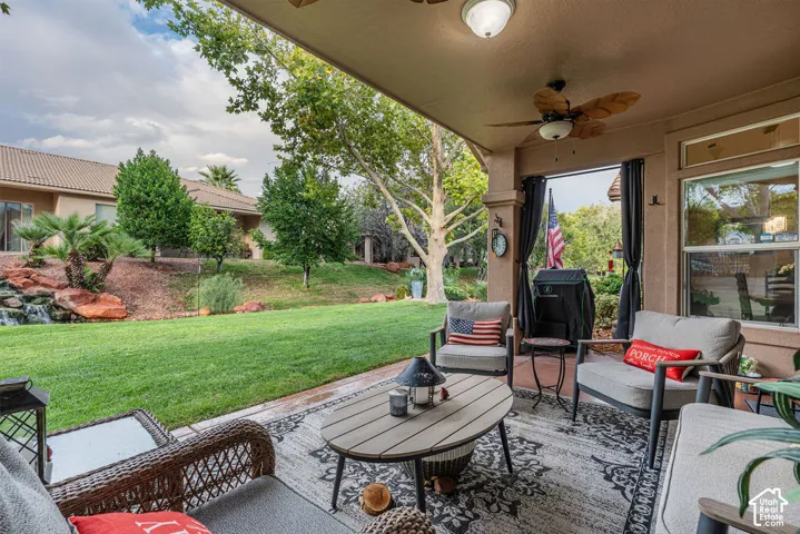 Covered patio featuring a ceiling fan and exterior lighting, overlooking a manicured lawn with mature trees and a stone-accented water feature