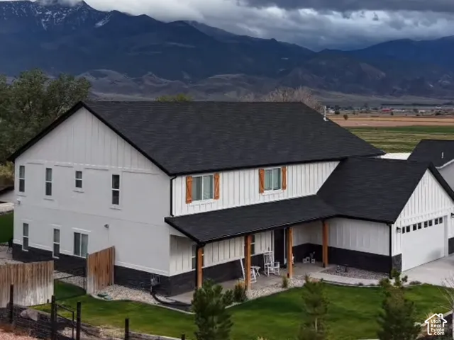 Multi-story residence featuring white board-and-batten siding, contrasting dark lower masonry, wood-accented shutters, a covered porch with wood support posts, and mountain-range scenery
