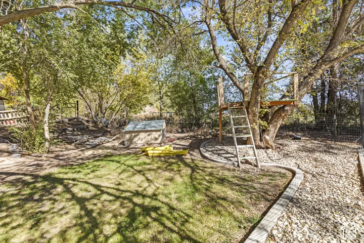 Private backyard featuring mature trees, a grass lawn, decorative gravel landscaping with concrete edging, and an elevated wooden platform structure with a fixed ladder