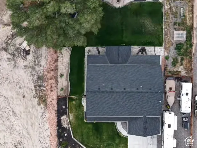 Aerial perspective featuring a shingled roof, a landscaped lawn, defined garden beds, stone-bordered pathways, and a gravel parking area