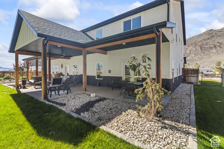 Covered rear patio with timber support beams, stone-veneer siding accents, manicured lawn, stone landscaping border, and mountain backdrop