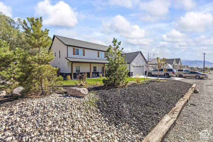 Two-story farmhouse-style exterior featuring board and batten siding, dark shingled roof, covered front porch with timber support posts, multi-car garage, and landscaped yard with decorative stone and timber retaining walls
