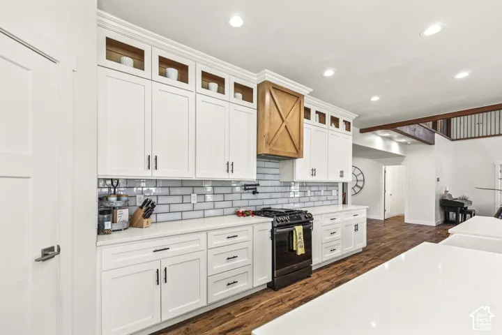 Kitchen featuring floor-to-ceiling white shaker cabinetry with glass-front display uppers, wood-tone range hood enclosure, light-colored stone-finish countertops, subway tile backsplash, and dark wood-finish flooring