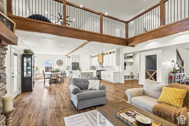Open-concept living space featuring wood-finish flooring, a double-height ceiling, a wood-beam balcony with metal railings, recessed lighting, and a kitchen equipped with white cabinetry and a center island