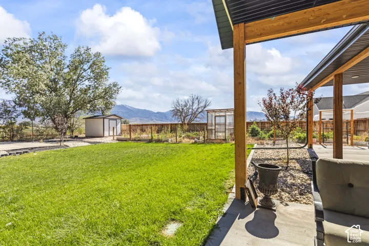 Covered concrete patio with timber support posts, manicured lawn, detached storage shed, raised garden beds, and mountain scenery