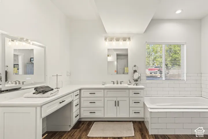 Bathroom featuring a double-sink vanity with white cabinetry, light-toned countertops, subway-tile tub surround, and wood-finish flooring