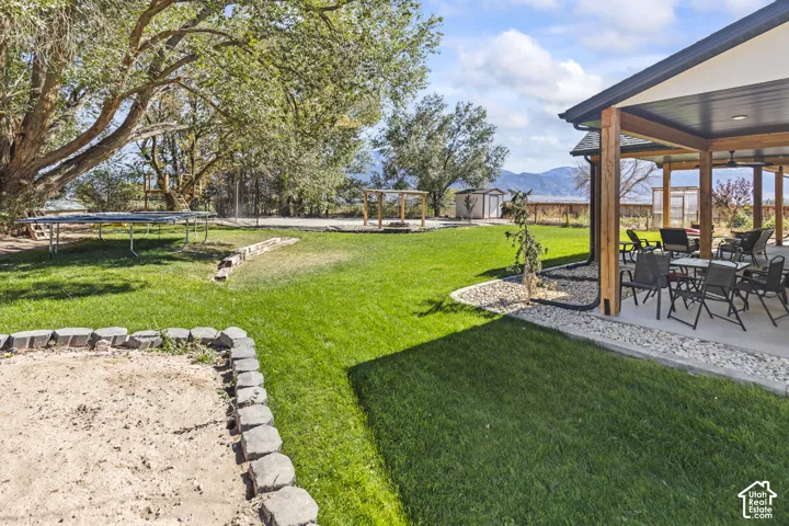 Expansive backyard featuring a covered concrete patio with timber support posts, a stone-bordered sand pit, manicured lawn, and mature trees