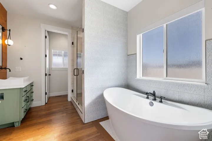 Freestanding soaking tub with matte black deck-mounted faucet, glass-enclosed shower with tiled surround, custom cabinetry vanity with vessel sink, and wood-finish flooring
