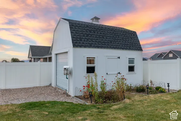 Detached outbuilding featuring white vertical siding, a gambrel roof with asphalt shingles, a roll-up garage door, and dual windows
