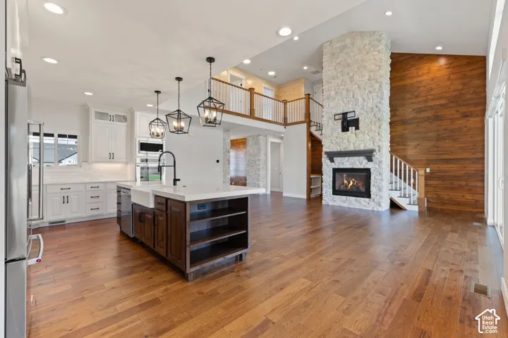 Open-concept kitchen and living area featuring hardwood flooring, a full-height stone fireplace, a wood-paneled accent wall, a kitchen island with a farmhouse sink, and three pendant lights