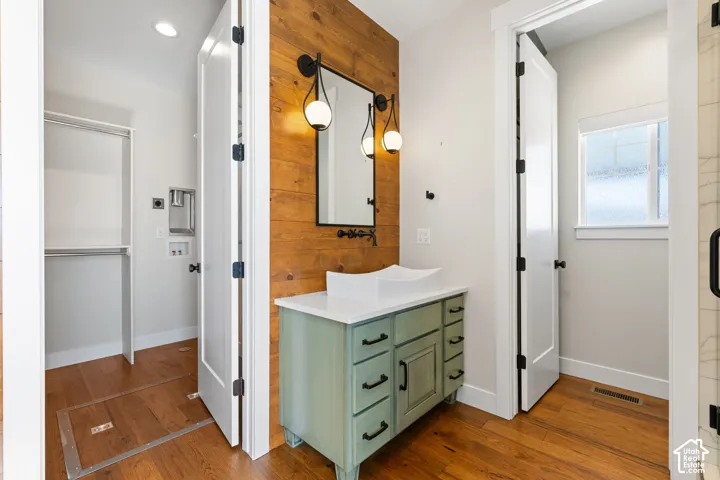Green vanity with a white countertop and vessel sink, wood-paneled accent wall, dual globe wall-mounted light fixtures, and wood-finish flooring