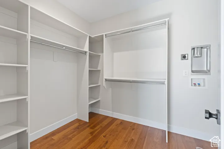 Primary ensuite closet with built-in white shelving, multi-level hanging rods, dedicated hookups for stacked laundry unit, and hardwood flooring