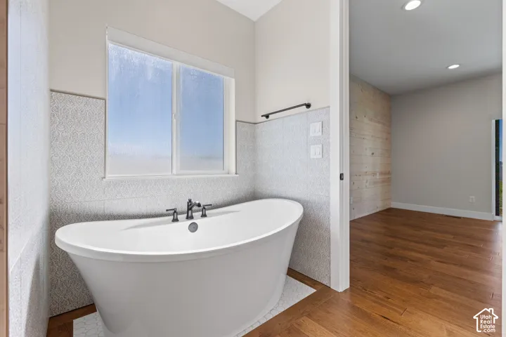 Freestanding soaking tub with a deck-mounted faucet, patterned tile surround, wood-finish flooring, and a decorative wood-plank accent wall