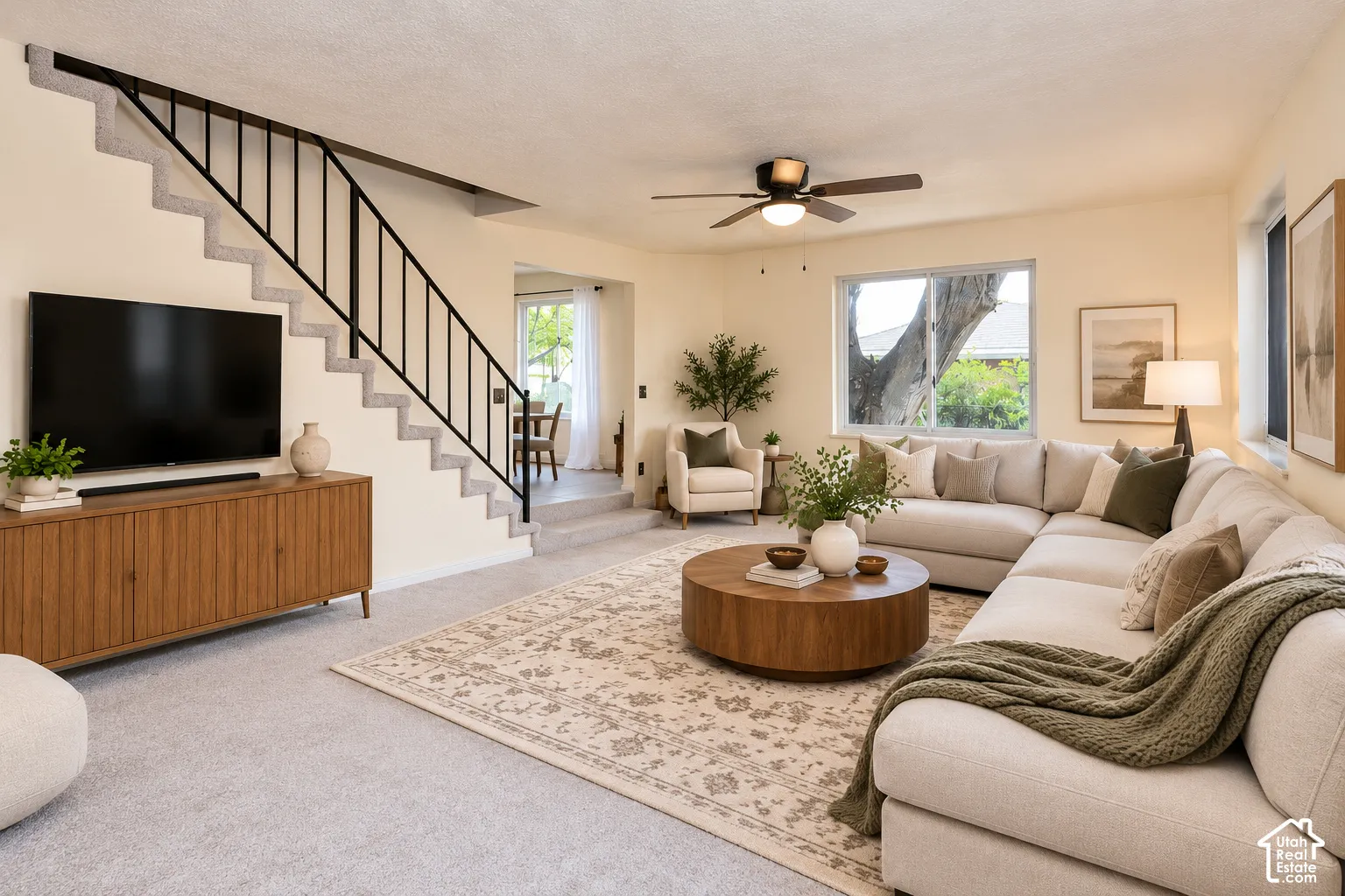 Living area featuring a staircase with a black metal railing, neutral carpeting, ceiling fan with integrated lighting, and large windows