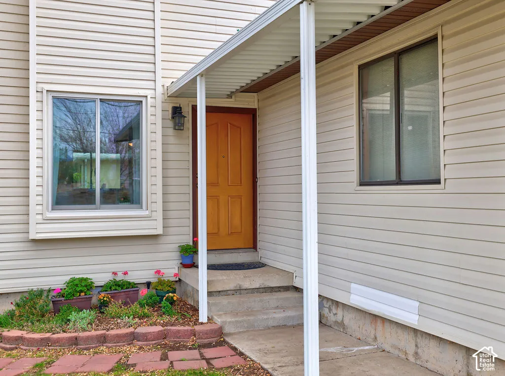 Exterior entrance featuring beige horizontal siding, a covered porch with metal support columns, a solid wood-finish entry door, and a tiered concrete stoop with stone-bordered garden bed