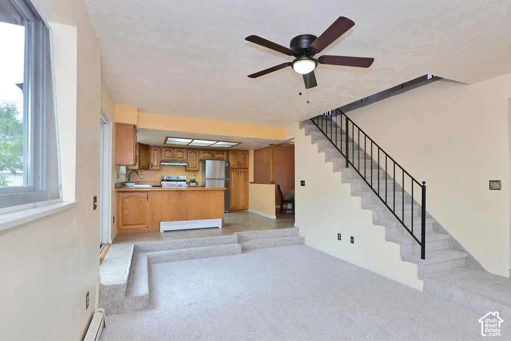 Sunken living area with carpeted tiered steps, open-concept kitchen featuring wood cabinetry, stainless steel appliances, and integrated lighting, paired with a staircase featuring a metal railing