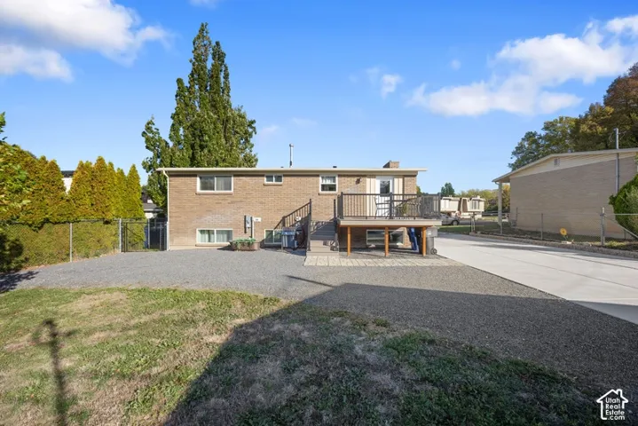 Brick exterior featuring an elevated deck with metal railing, gravel parking area, concrete driveway, and chain-link perimeter fencing