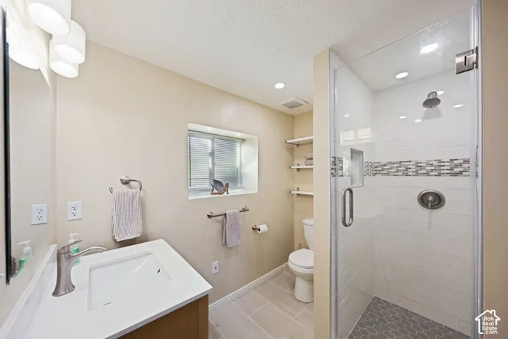 Bathroom featuring a vanity with a white rectangular sink, a glass-enclosed shower with tiled walls and a mosaic accent border, hex-tile shower flooring, and wall-mounted shelving