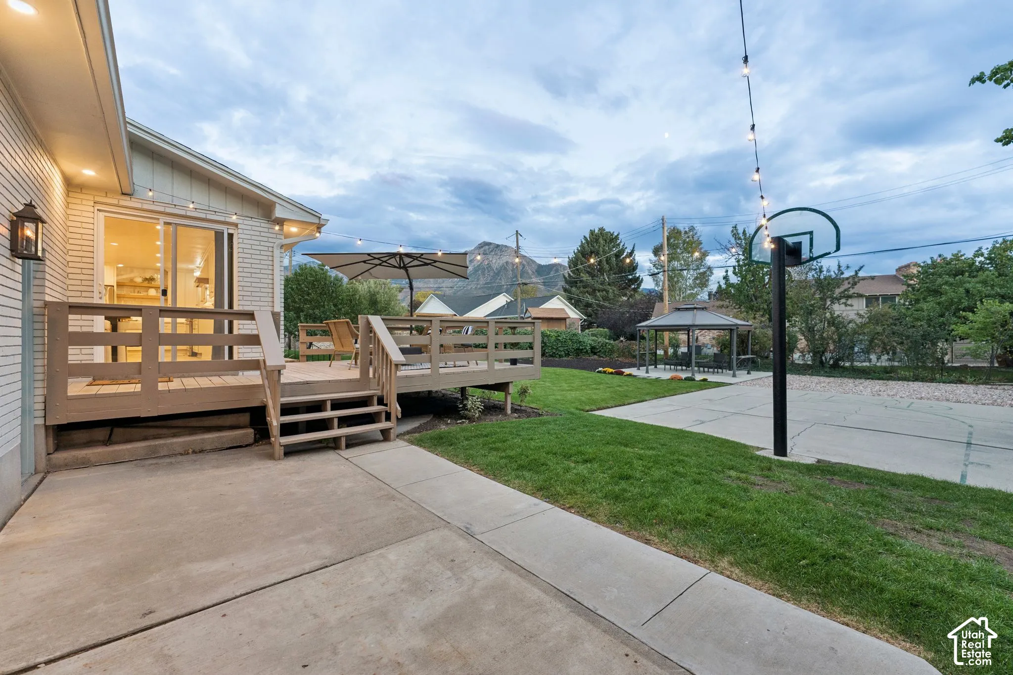 Rear exterior featuring a raised wood deck, concrete patio, built-in basketball hoop, and manicured lawn with mountain backdrop