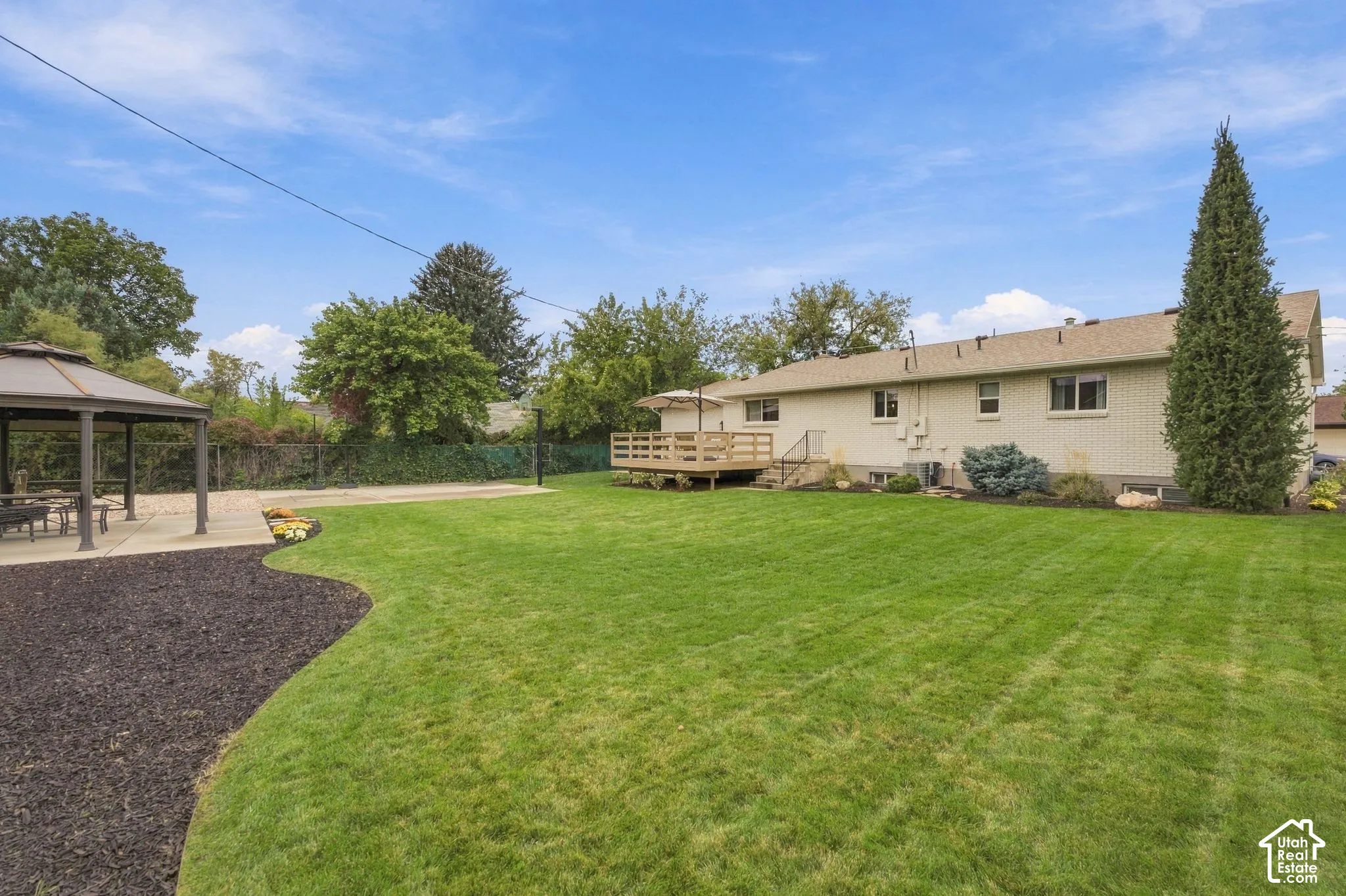 Expansive backyard featuring a manicured lawn, detached hardtop gazebo, raised wood deck, and perimeter chain-link fencing