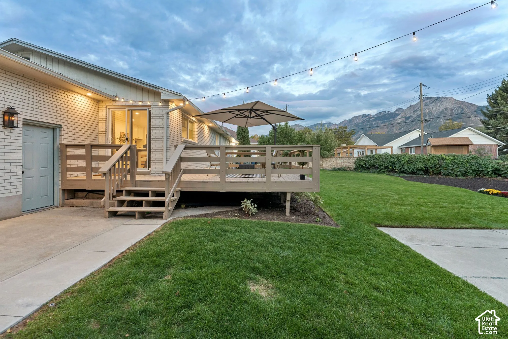 Raised wood deck with integrated railings, brick exterior siding, sliding glass door entry, concrete patio, and mountain backdrop
