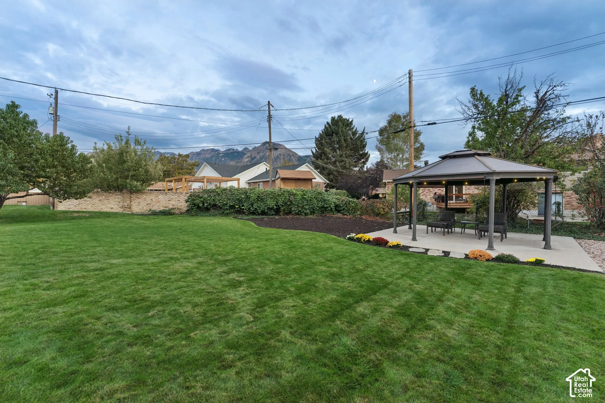 Expansive backyard featuring a manicured lawn, stone retaining wall, paved patio area with a gazebo, and mountain backdrop