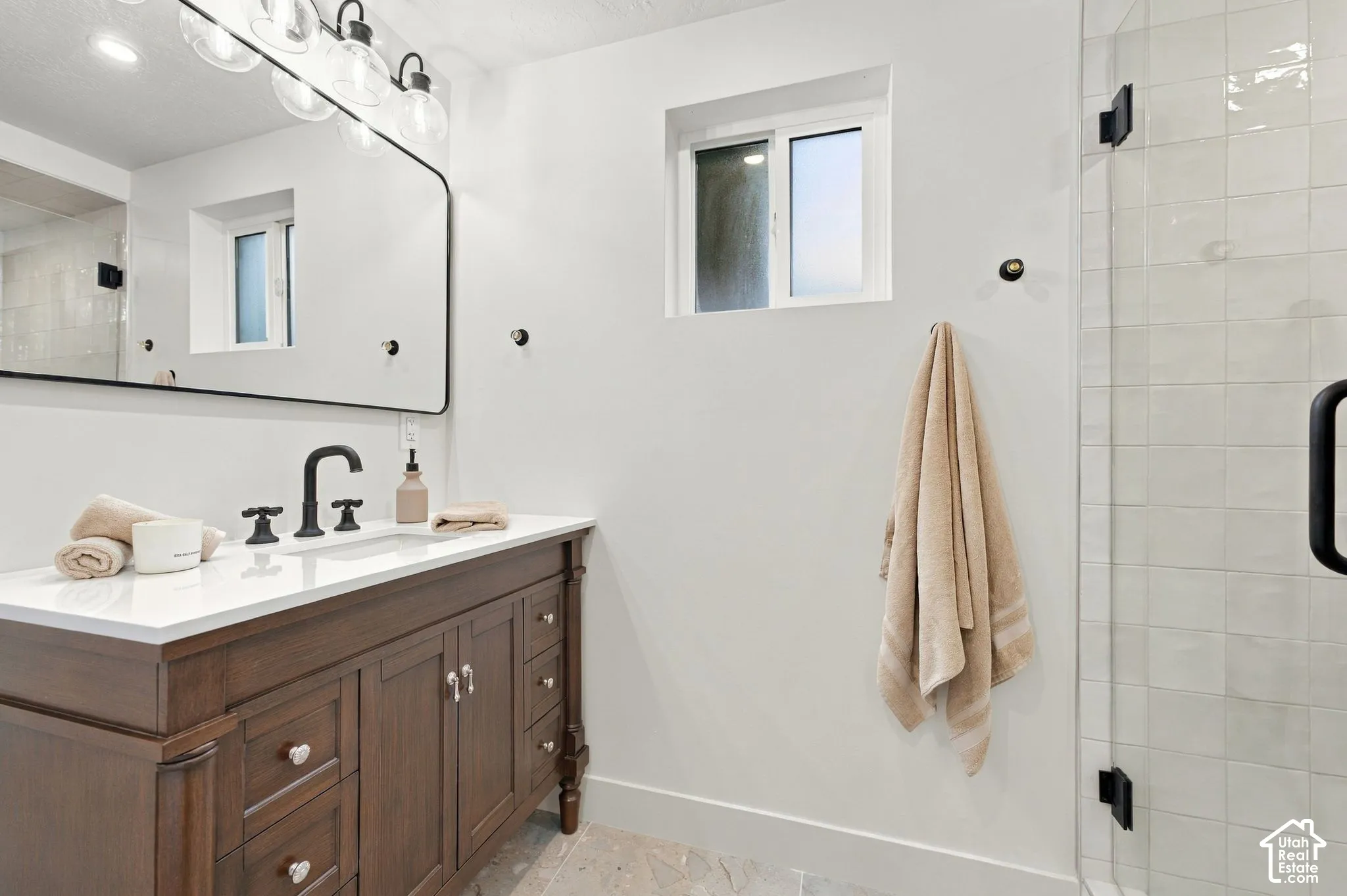 Wood-finish cabinetry vanity with a white countertop and matte black faucet, glass-enclosed shower with tiled surround, and large wall-mounted mirror with integrated overhead lighting