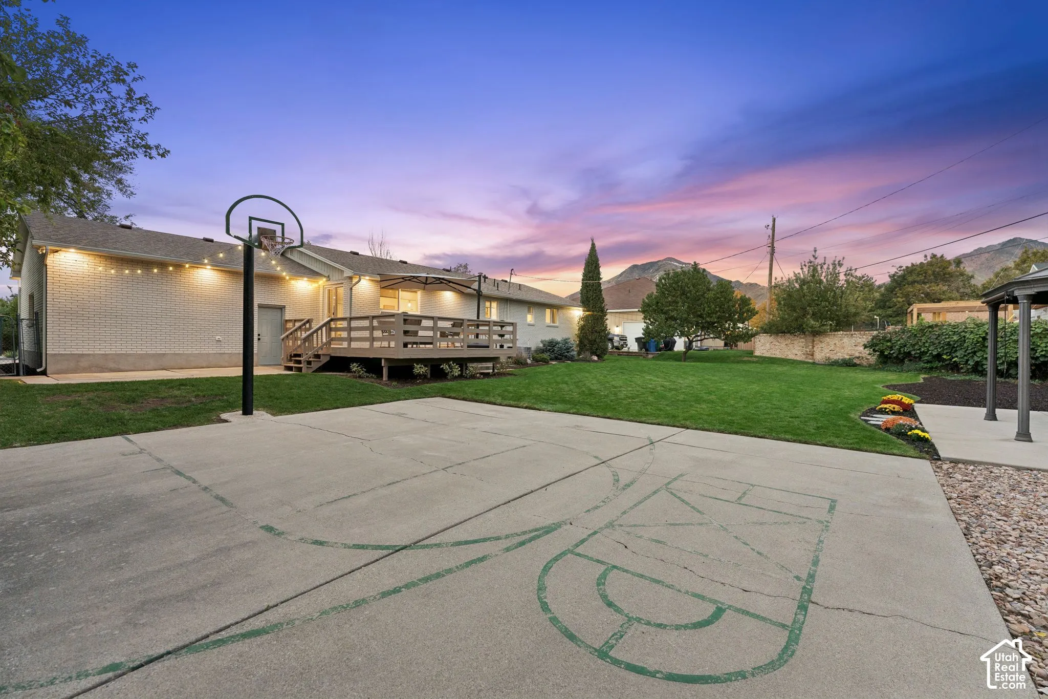 Backyard featuring a concrete basketball court, wooden deck, manicured lawn, stone retaining wall, and mountain backdrop