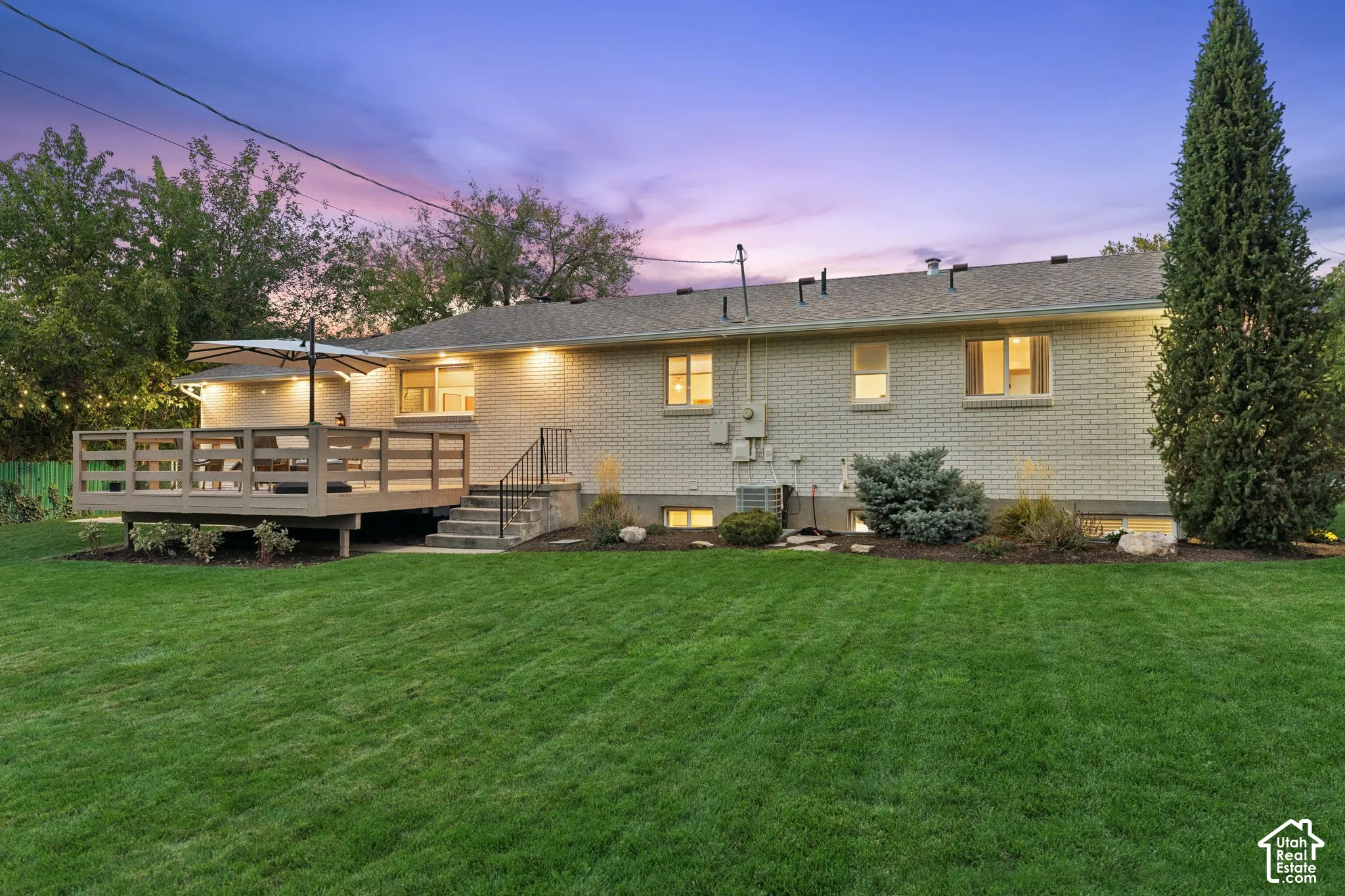 Exterior brick facade featuring a raised wooden deck, staircase with metal railing, manicured lawn, and mature landscaping