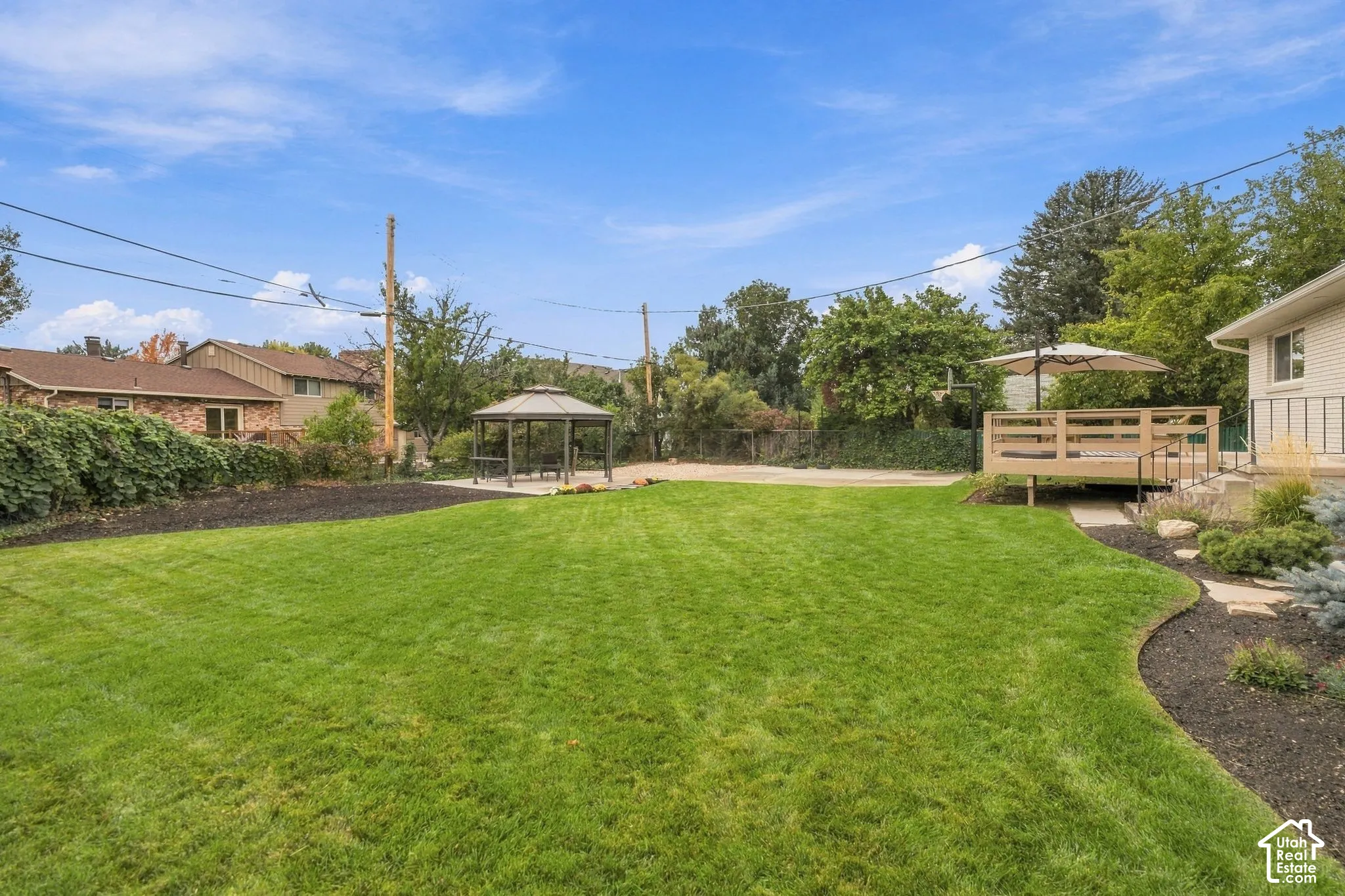 Expansive backyard featuring a manicured lawn, tiered wooden deck, permanent metal gazebo, and decorative stone landscaping beds