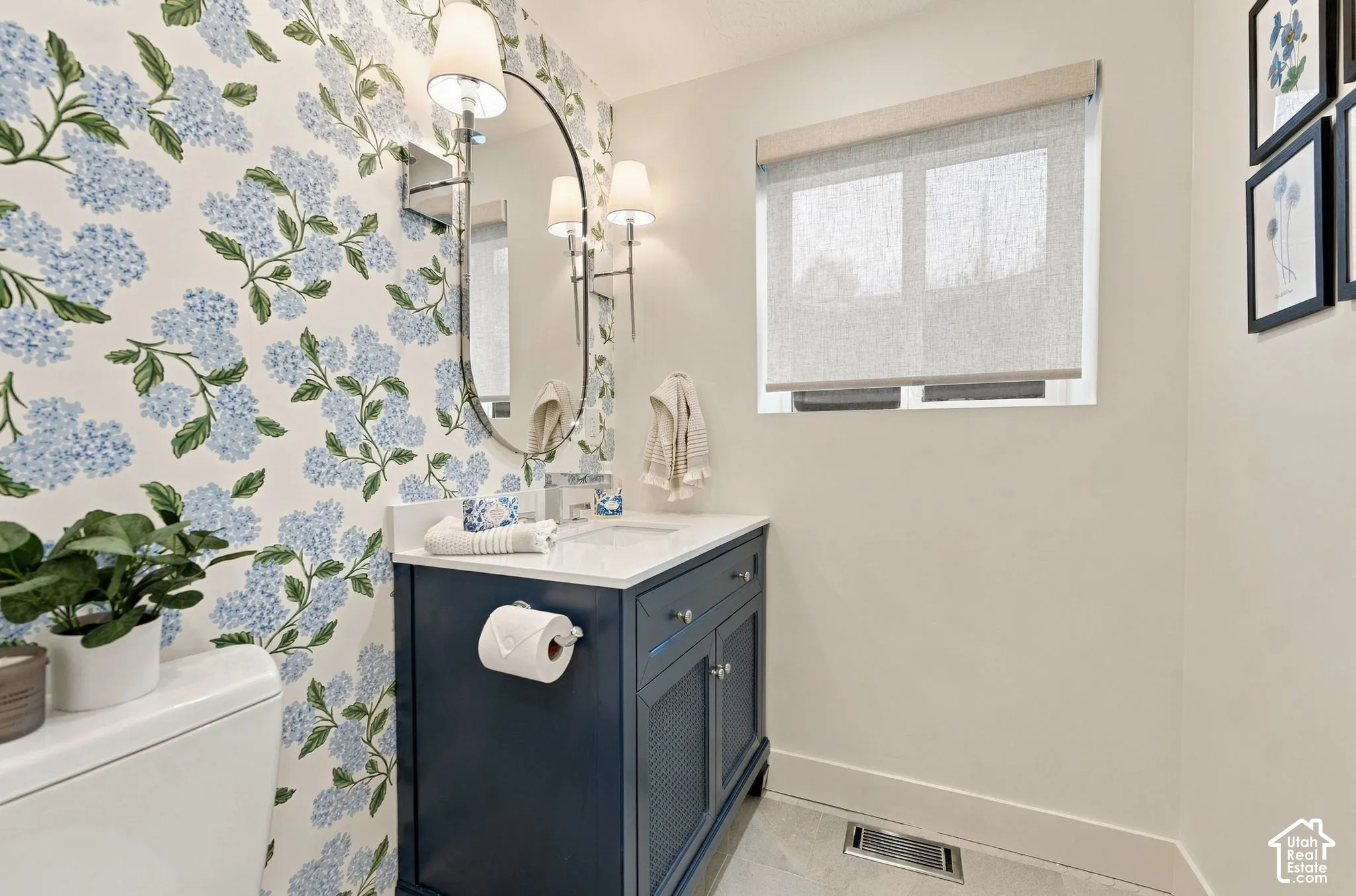 Custom navy blue vanity with a stone countertop, oval wall-mounted mirror, dual polished metal light fixtures, and floral-patterned wall covering